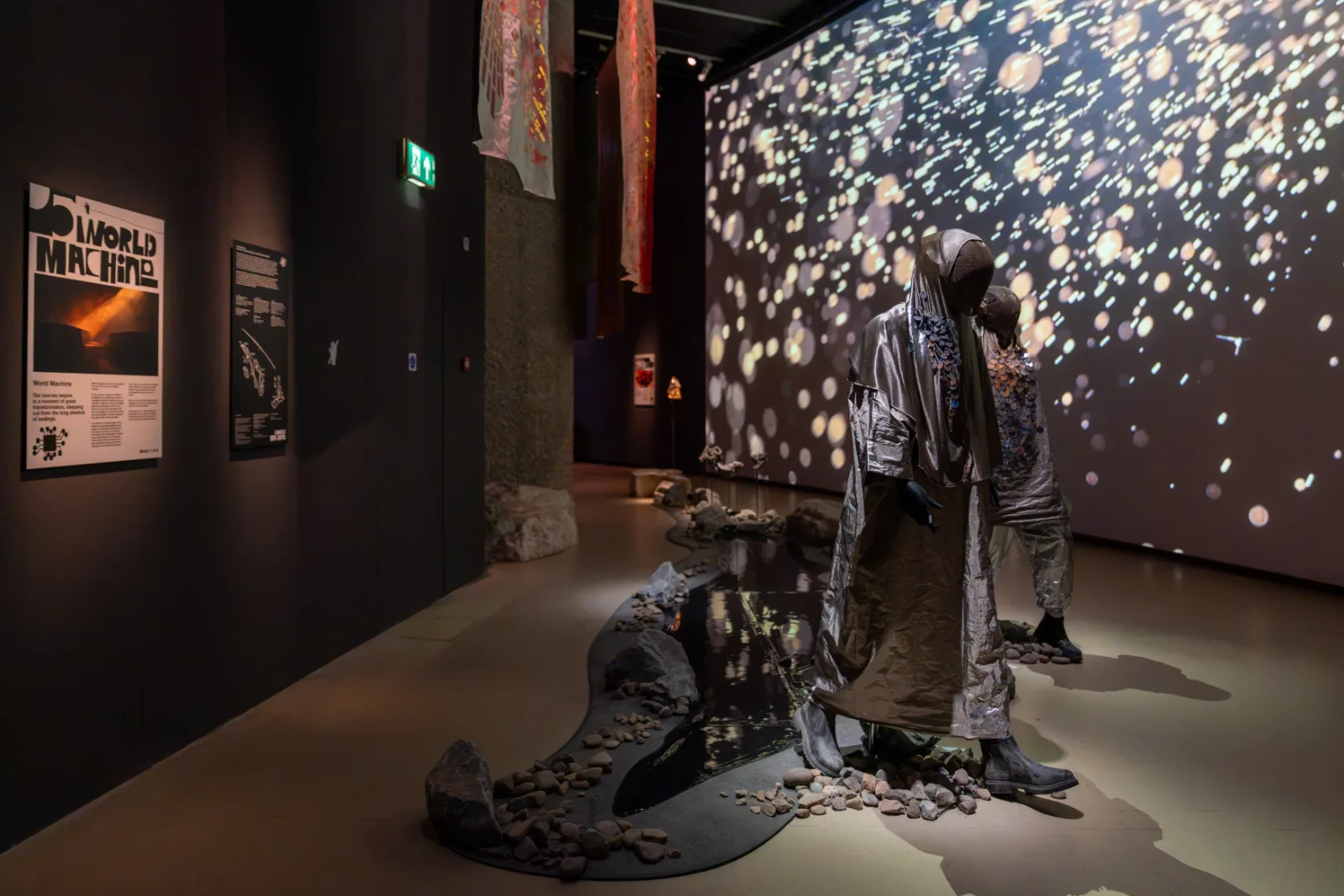 Two costumed figures standing beside a channel of water in front of a large projection of scattered light, World Machine, In Other Worlds, Barbican Centre, London, 2026