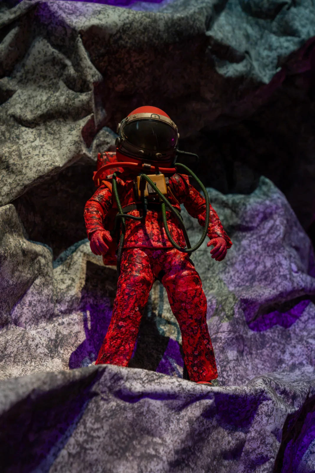 A small figure in a red floral pressure suit and orange helmet standing among pale rock forms lit in purple, In Other Worlds, Barbican Centre, London, 2026