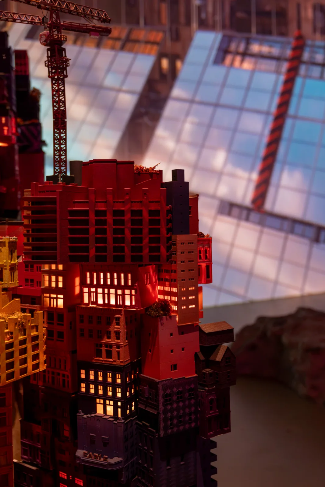 A dense stack of miniature buildings with lit windows and a construction crane set against a large projection, In Other Worlds, Barbican Centre, London, 2026