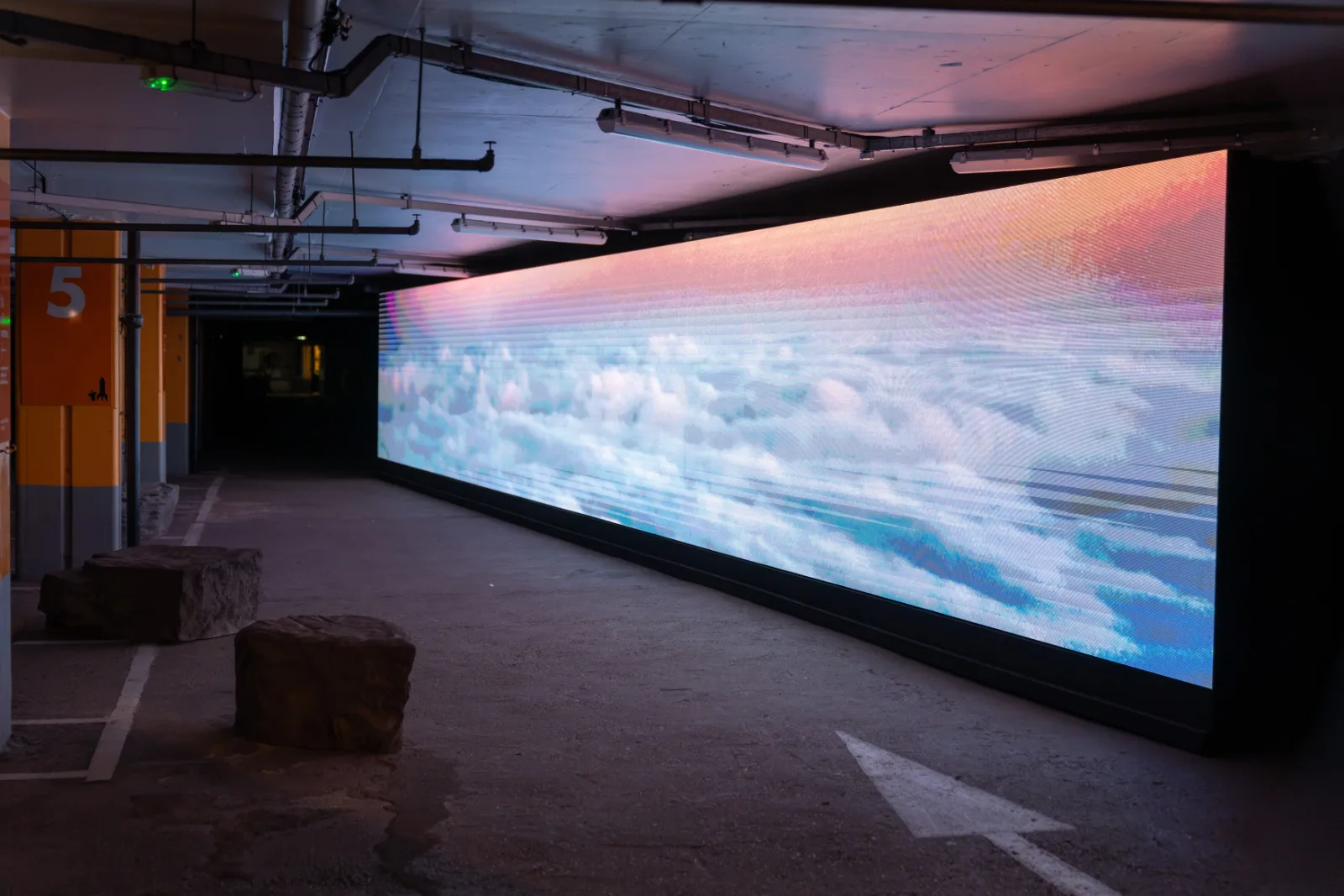 A long low LED screen showing clouds and sky installed along a car park level marked 5, In Other Worlds, Barbican Centre, London, 2026