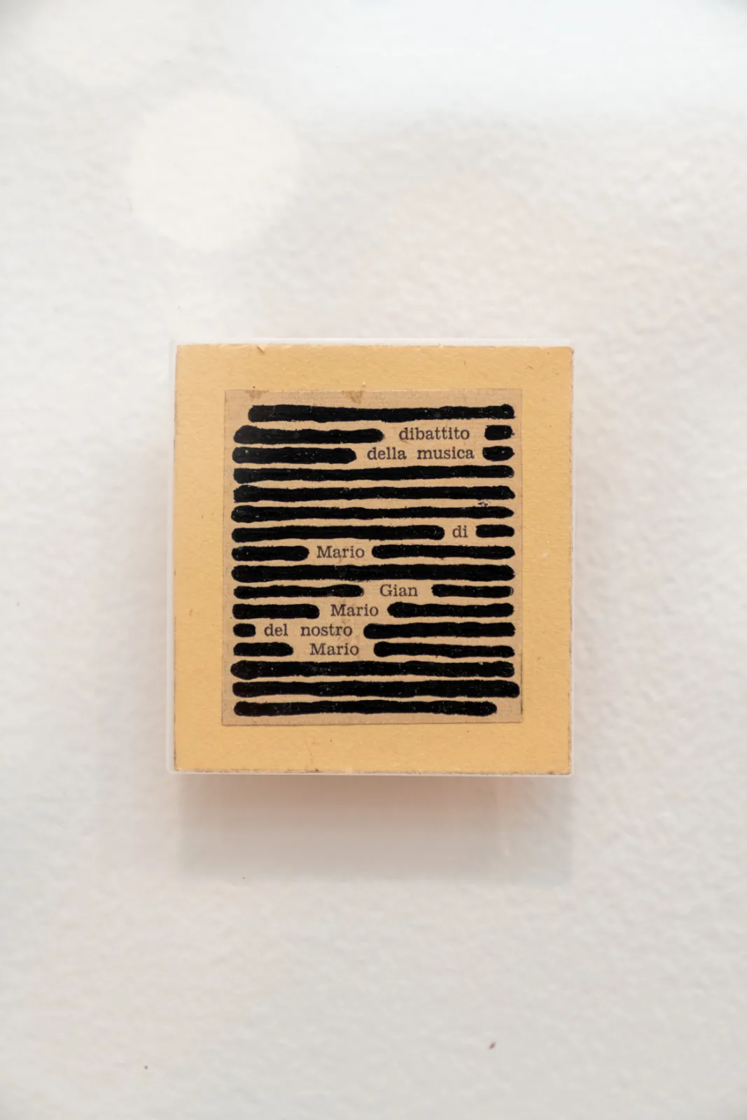 A small square of newsprint sealed in a plexiglass block, almost entirely covered in thick black ink strokes, with the words dibattito, della musica and the first name Mario surviving three times