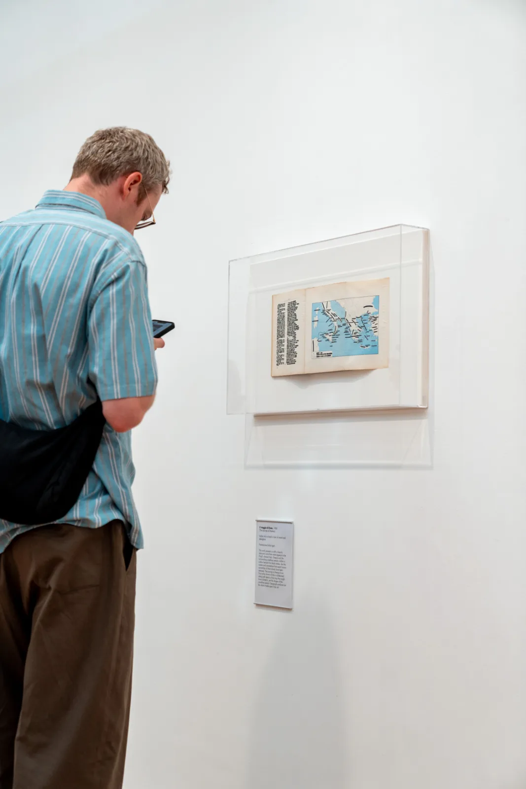A visitor seen from behind looking at a small wall case holding an open book, its left page a canto struck through with black ink and its right page a blue map of the Mediterranean with the coastlines visible and the place names erased