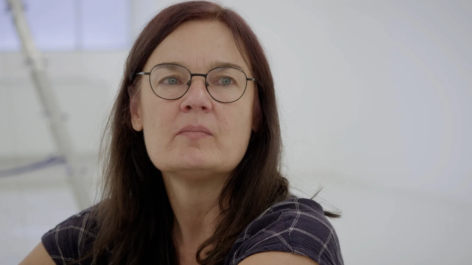 Close portrait of a woman with long dark hair and round metal glasses, wearing a dark checked shirt, looking past the camera in a white room.