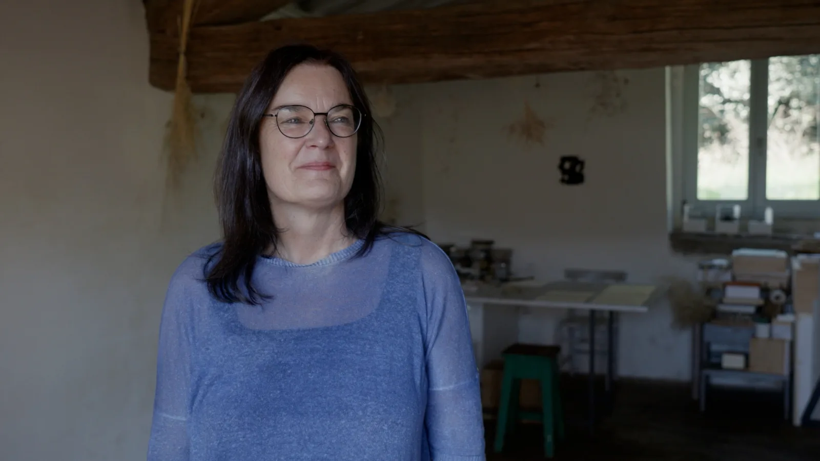 A woman with dark hair and glasses, in a blue sweater, smiles slightly in a studio with a dark wooden beam, a work table, boxes and a window onto greenery.