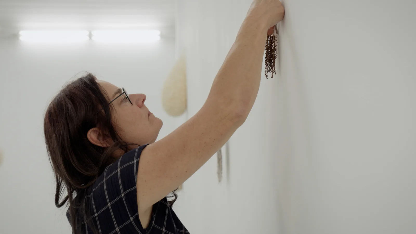 In profile, a woman with glasses stretches her arm up to fix a hanging work of dark seed strands to a white wall; a pale teardrop-shaped work hangs further along.