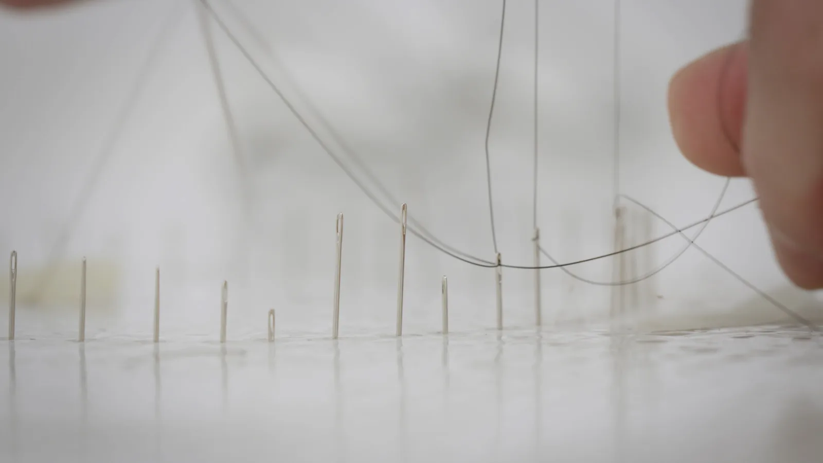 Low view of sewing needles of different heights standing upright in a white surface, with strands of horsehair threaded and stretched between them and a fingertip in the foreground.