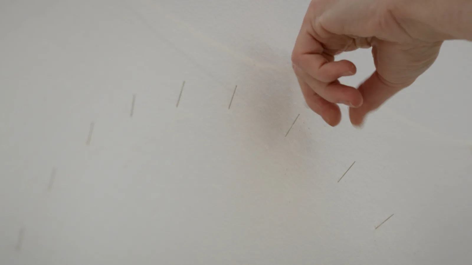 A hand reaches toward a white wall where a curved row of thin needles has been set at regular intervals.