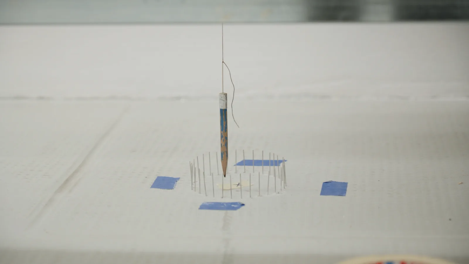 A worn blue pencil hangs from a thread, its tip just above a ring of pins on a white board, with four strips of blue tape around it.