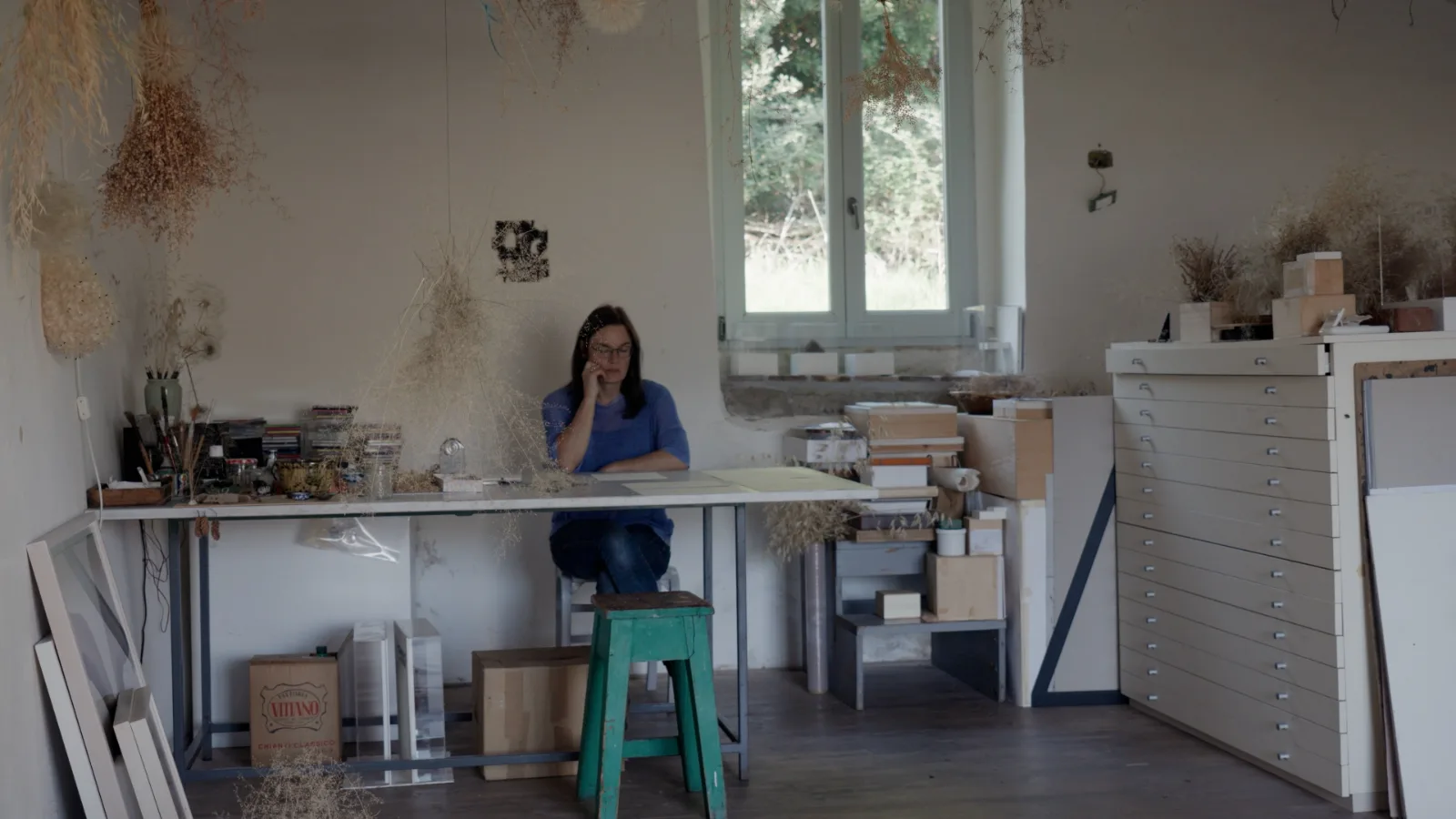 Wide view of a studio with bunches of dried grasses and seed heads hanging from the ceiling; a woman sits at a long table under a window, with a green stool in front and a tall white plan chest on the right.