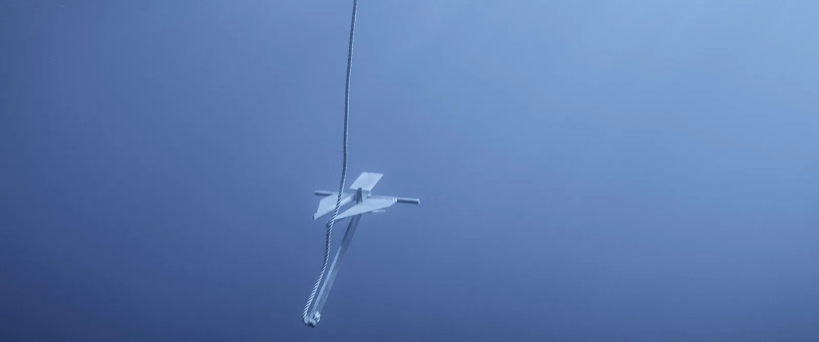 A boat anchor suspended in deep blue water on a rope disappearing upward, still from She Flickered In and Out of History