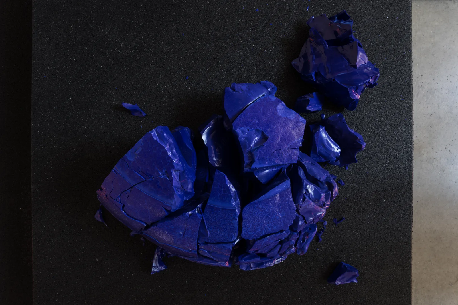 A cracked mass of deep blue cotisso glass seen from above on black rubber, its fragments scattered like a coastline