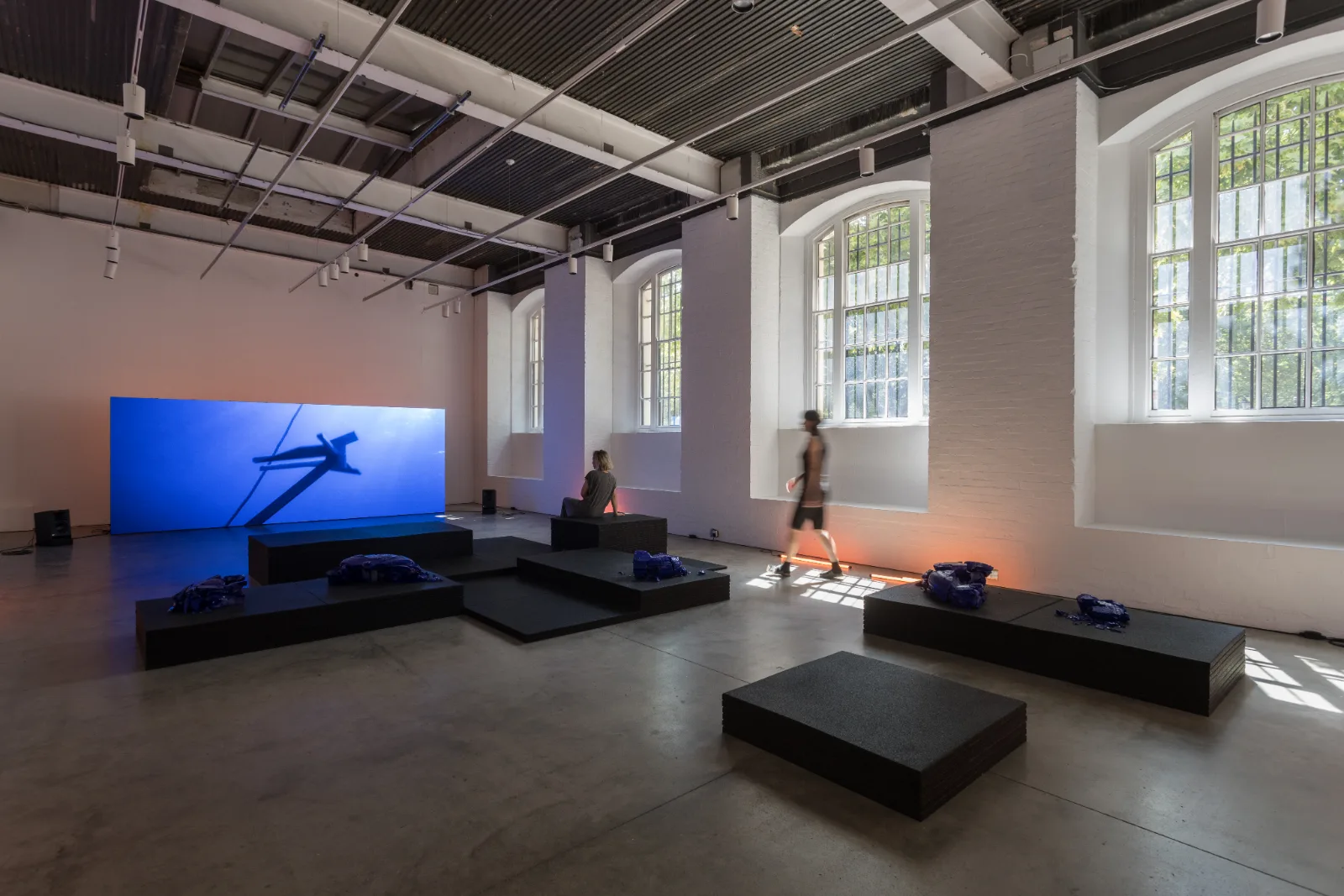 Installation view of She Flickered In and Out of History at the ICA London, with a free-standing LED screen and black rubber platforms in a daylit gallery
