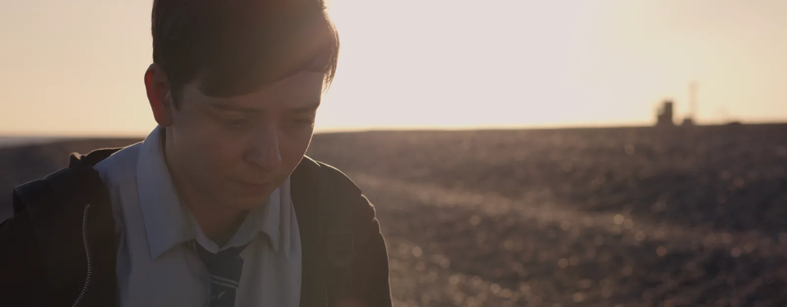 A boy in a school uniform looks down, backlit by a low sun, on a shingle landscape