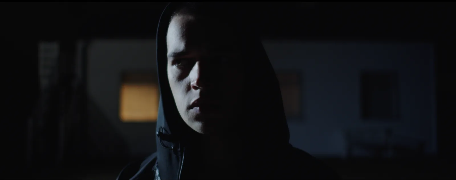 A young man in a dark hood, half his face in shadow, in a dimly lit room