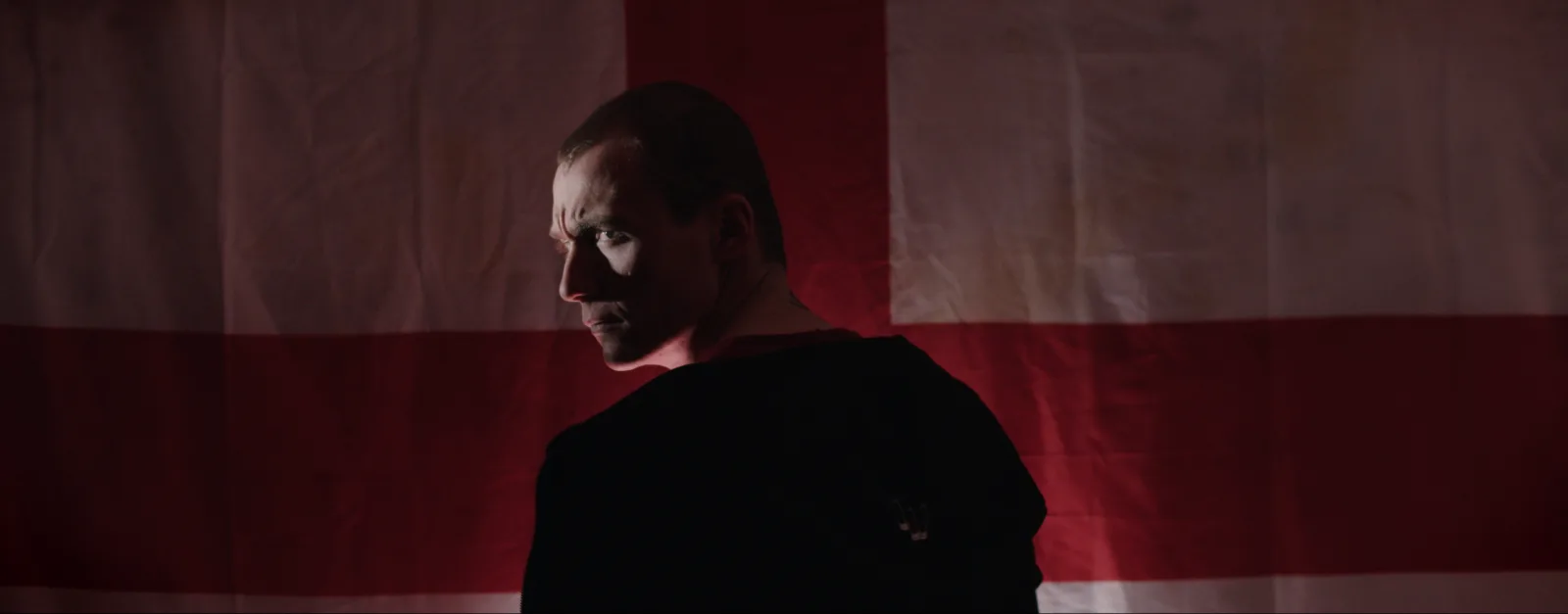A man with a shaved head looks over his shoulder in front of a St George Cross flag, his face half in shadow