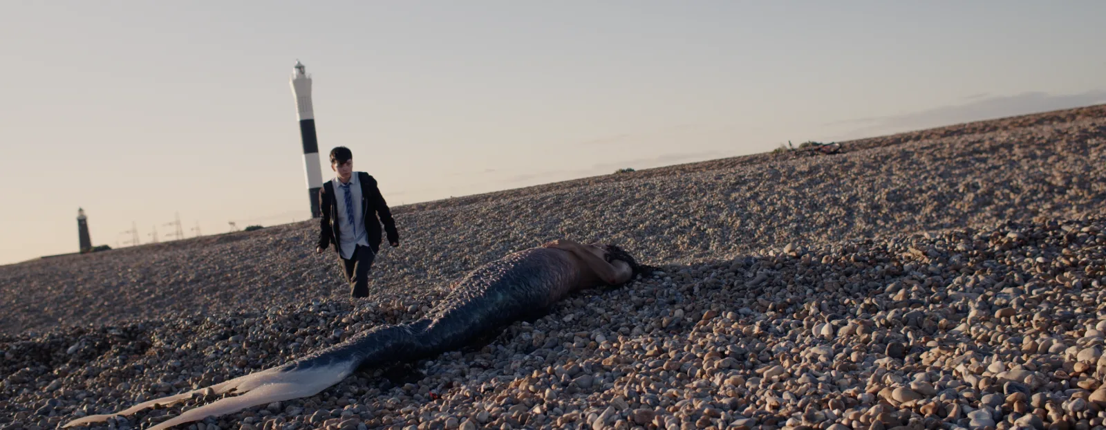 A boy in a school uniform walks across a shingle beach towards a mermaid lying on the stones, with a black and white lighthouse behind him
