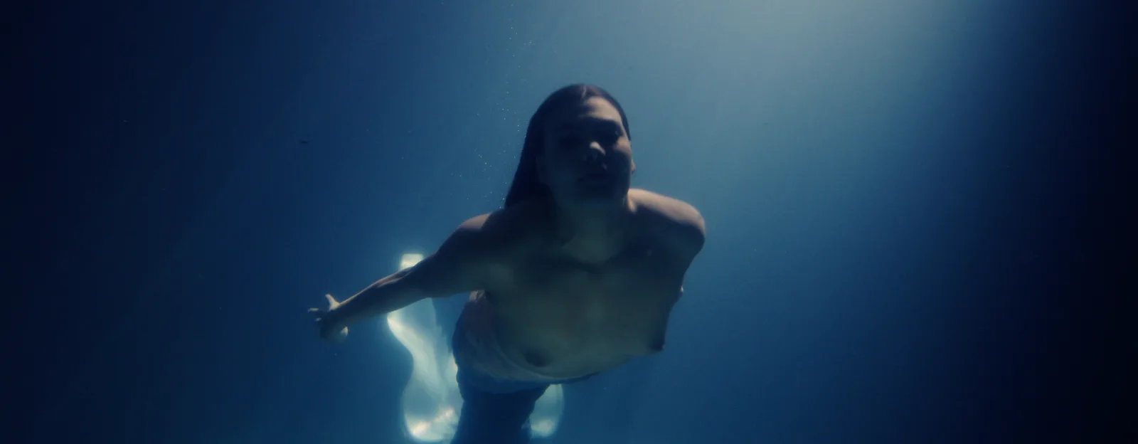 A mermaid with long dark hair swims towards the camera in deep blue water, one arm stretched out, her tail glowing pale behind her