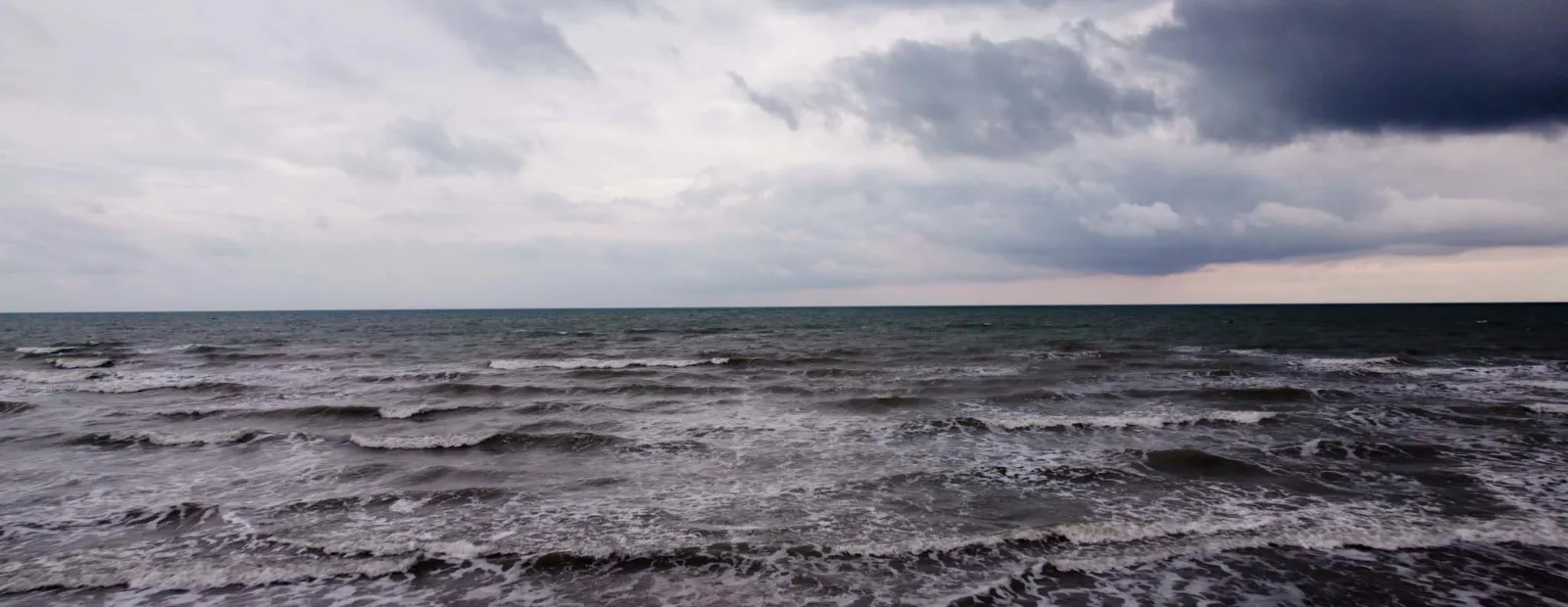 A grey sea under heavy storm clouds, waves breaking towards the shore and a straight horizon line