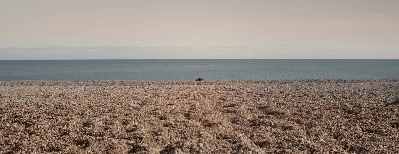 A wide expanse of pale shingle stretching to a calm grey sea, a tiny dark figure lying at the edge of the stones