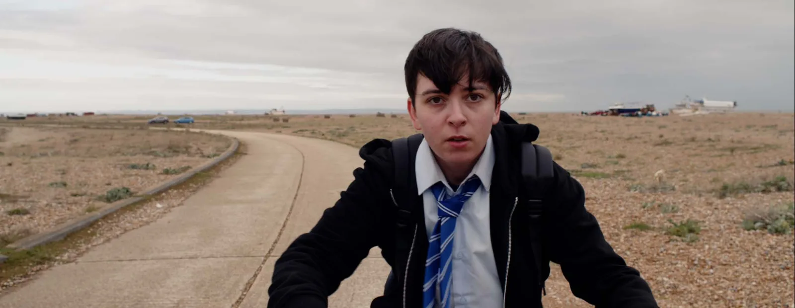 A boy in a school uniform and blue striped tie rides along a concrete road across the shingle, fishing boats and huts on the horizon