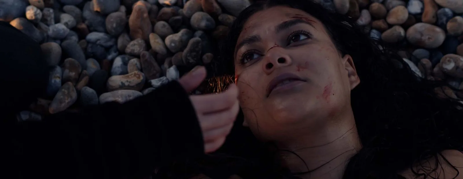 A hand in a dark sleeve reaches towards the wounded face of a woman with long dark hair lying on the pebbles