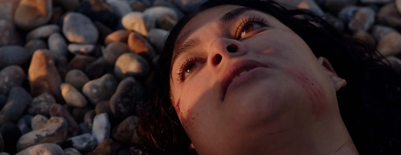 Close-up of a woman lying on the pebbles in warm evening light, her face scratched, her eyes turned up towards the sky