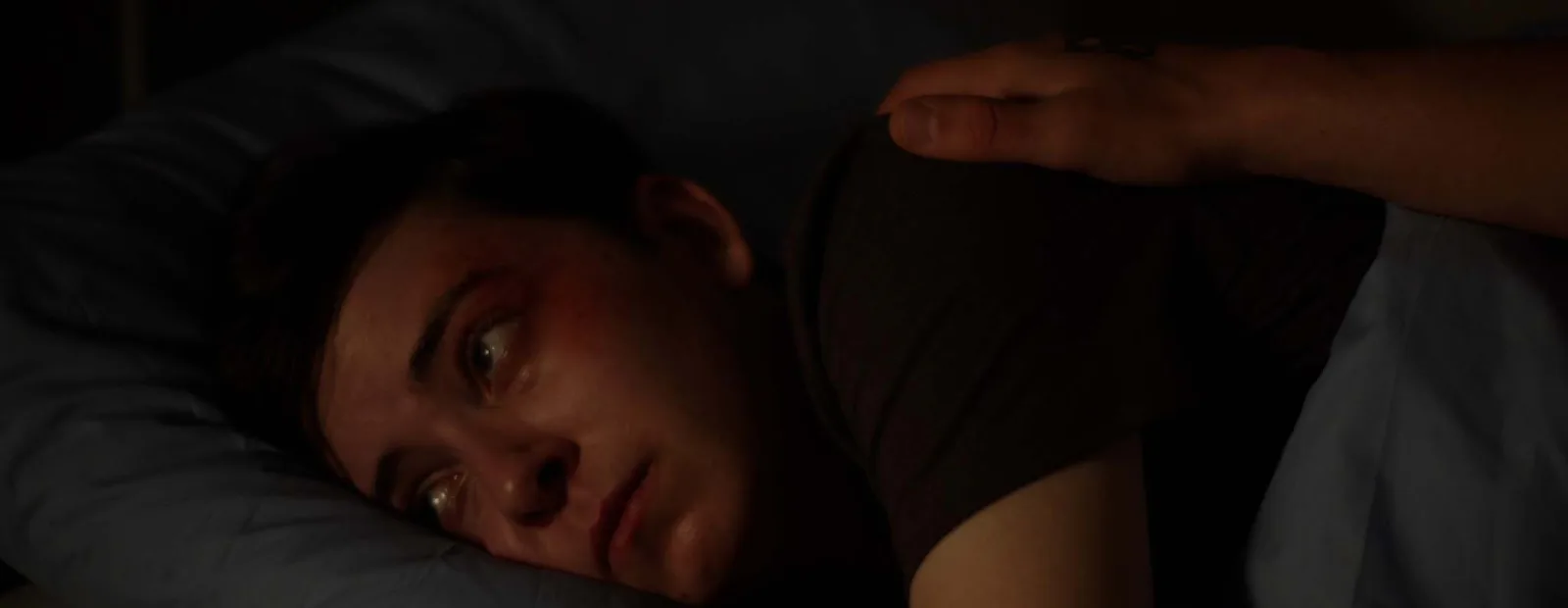 A boy lies awake in bed in the dark, a bruise around his eye, a hand resting on his shoulder