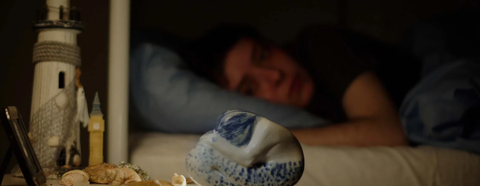 A small blue and white ceramic mermaid on a bedside table with shells and a toy lighthouse, a boy asleep out of focus behind it