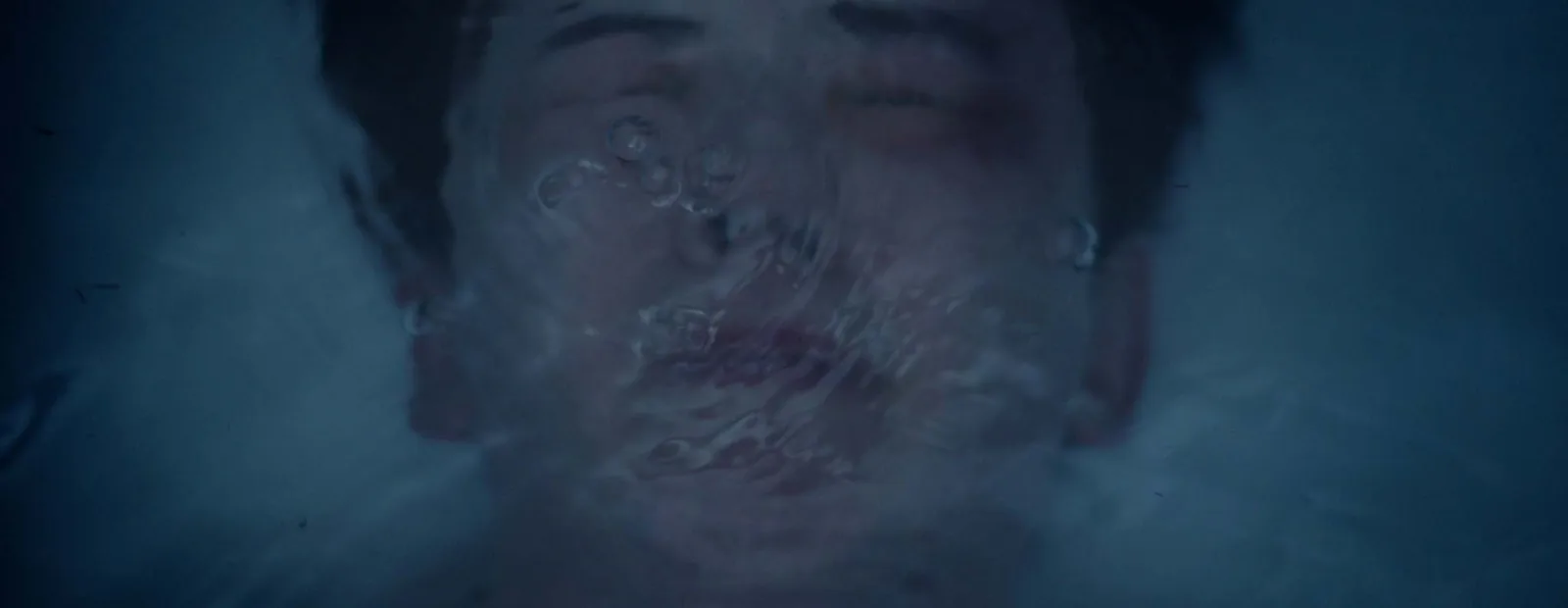 The face of a boy seen through the surface of the water, eyes closed, blurred by ripples and small bubbles