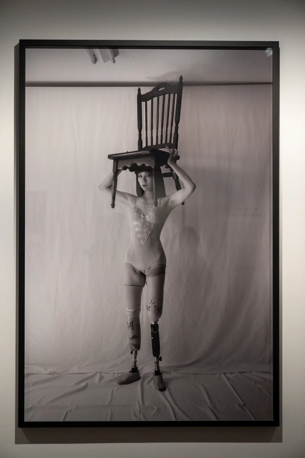 Framed black and white photograph of a woman with prosthetic legs, standing and holding a wooden chair above her head