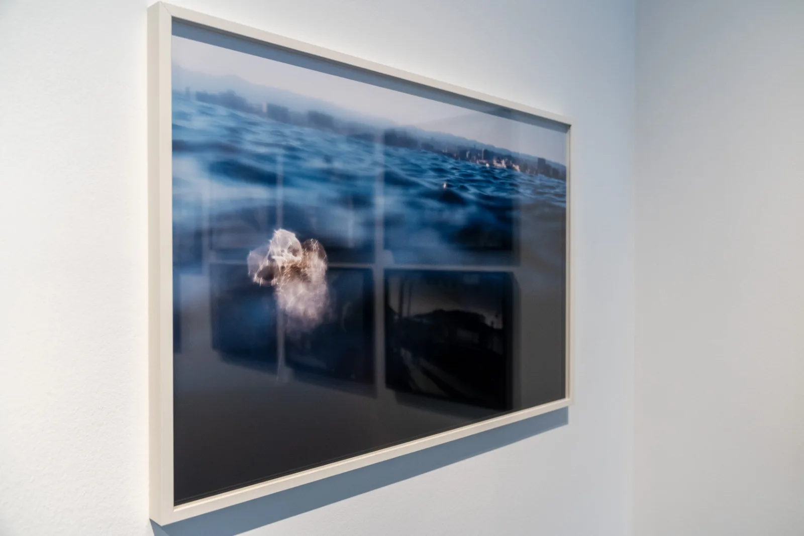 Framed colour photograph taken at the surface of the sea, with blue waves, a distant hazy coastline of buildings and a pale blurred shape in the water