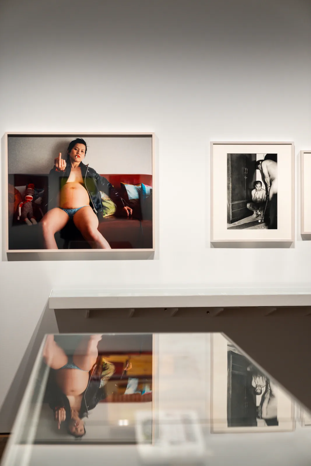Two framed photographs on a gallery wall reflected in a glass vitrine, on the left a pregnant woman in a leather jacket on a red sofa raising her middle finger, on the right a black and white image of a crouching woman and a standing man in a doorway