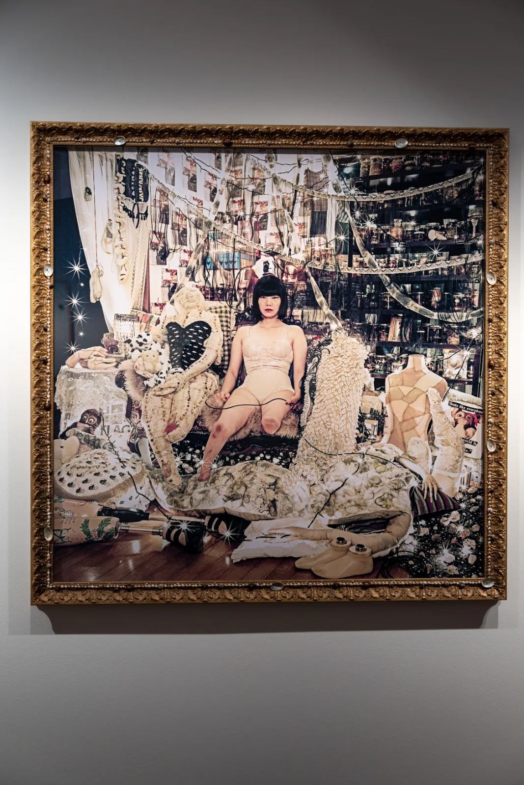 Photograph in an ornate gold frame of a seated woman surrounded by handmade textile objects, lace, shelves of jars and prosthetic legs on the floor