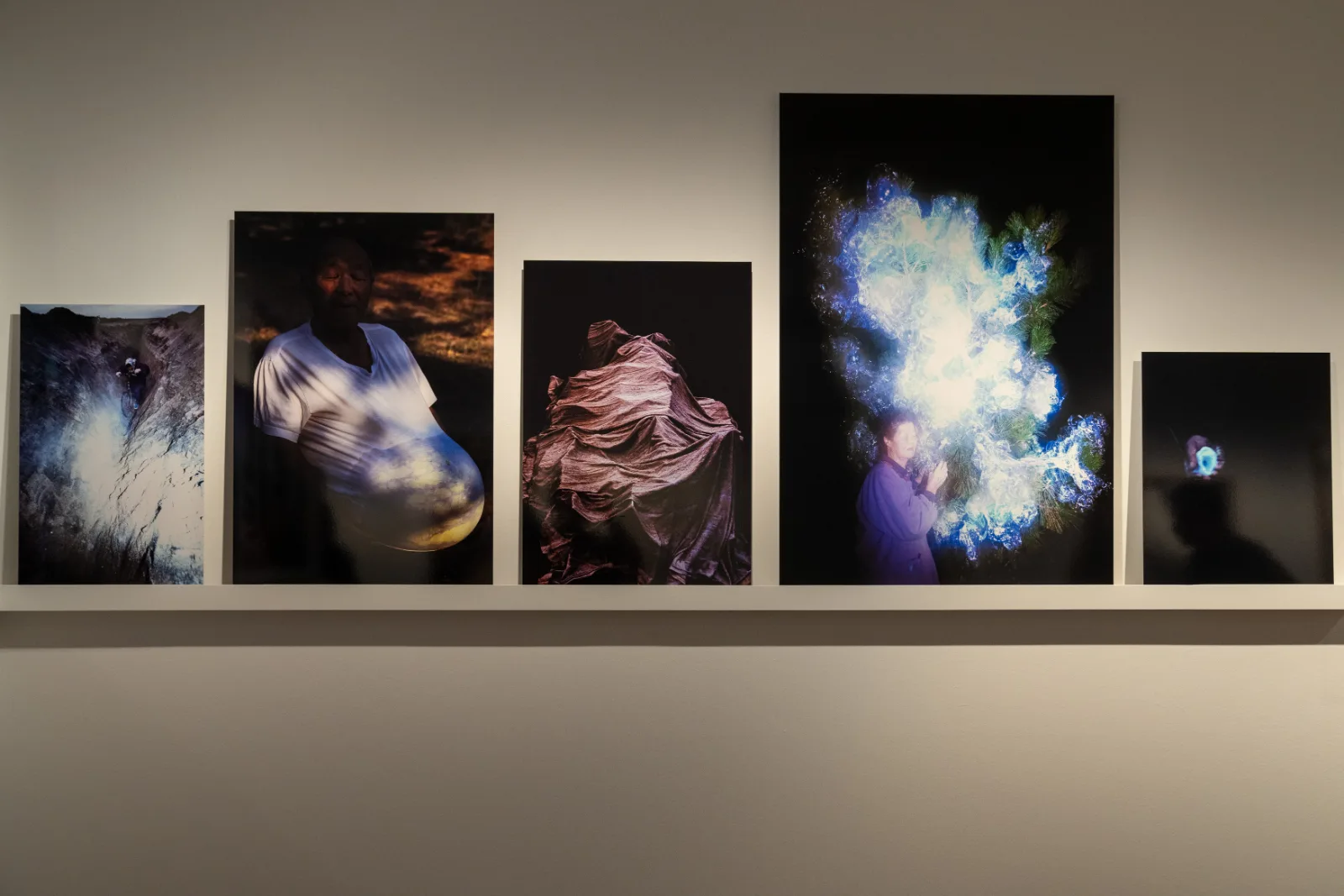 Five colour photographs of different sizes leaning on a shelf, among them an elderly man with a glowing sphere over his belly, a figure under a draped cloth and a woman beside a burst of blue light in pine branches