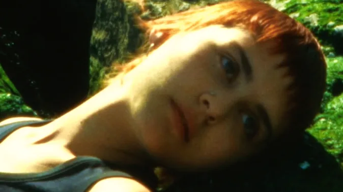 Close-up of Rory Bakker-Marshall lying on green seaweed, her red hair bright against the green, her eyes open towards the camera