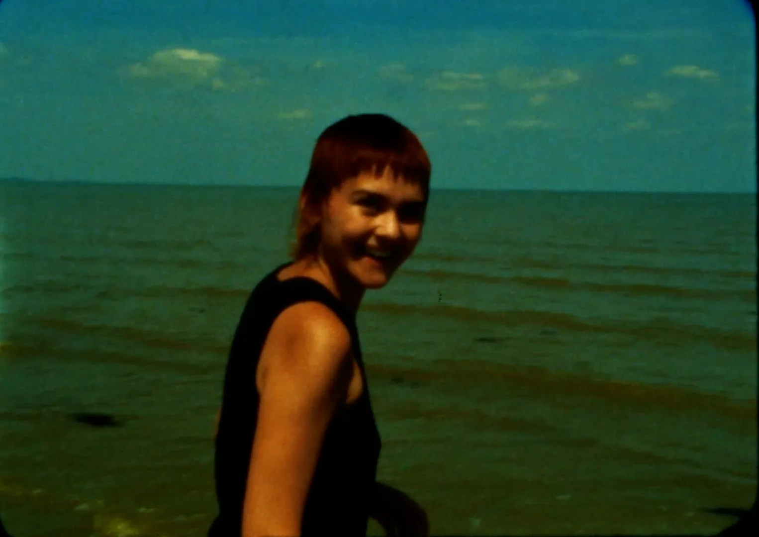 Rory Bakker-Marshall, with short red hair and a black sleeveless top, turns towards the camera and smiles, the sea and a blue sky behind her