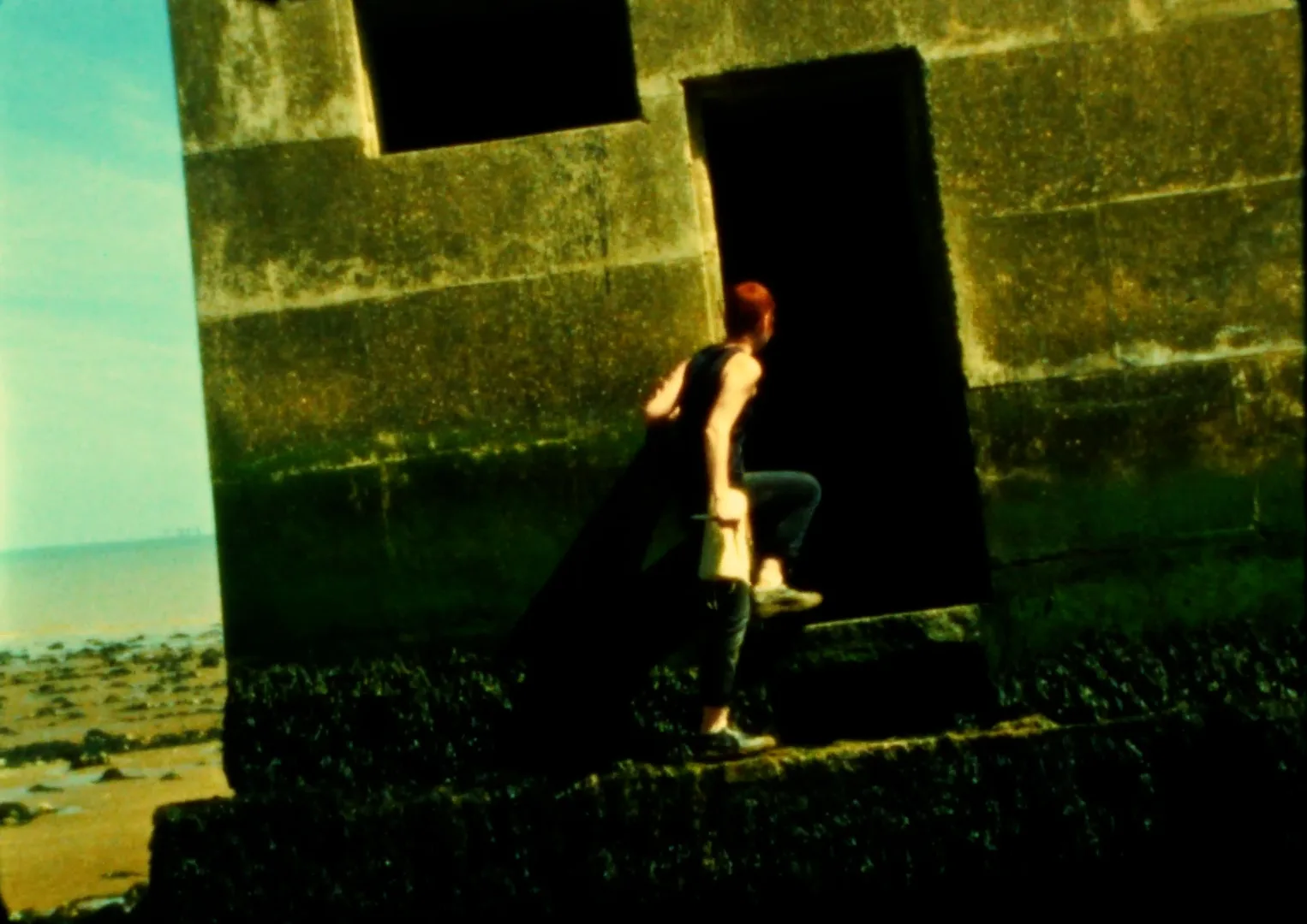 Rory Bakker-Marshall climbs into the dark doorway of a concrete pillbox covered in seaweed, a cloth bag in her hand, the beach at low tide behind her