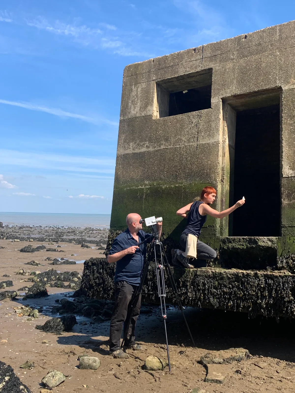 On a beach at low tide, a man films through a Super 8 camera on a tripod while Rory Bakker-Marshall kneels at the entrance of a concrete pillbox covered in seaweed, holding up a small white object