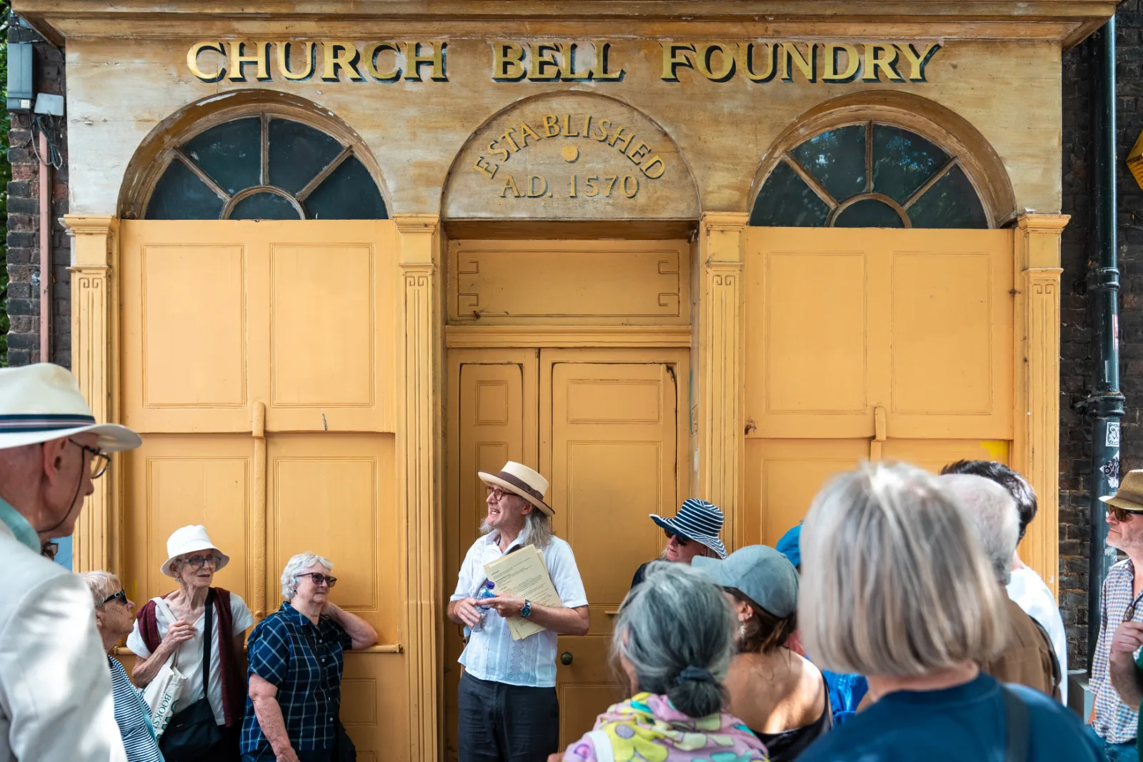 A walking group gathered outside the former Whitechapel Bell Foundry, its yellow facade lettered Church Bell Foundry, Established A.D. 1570