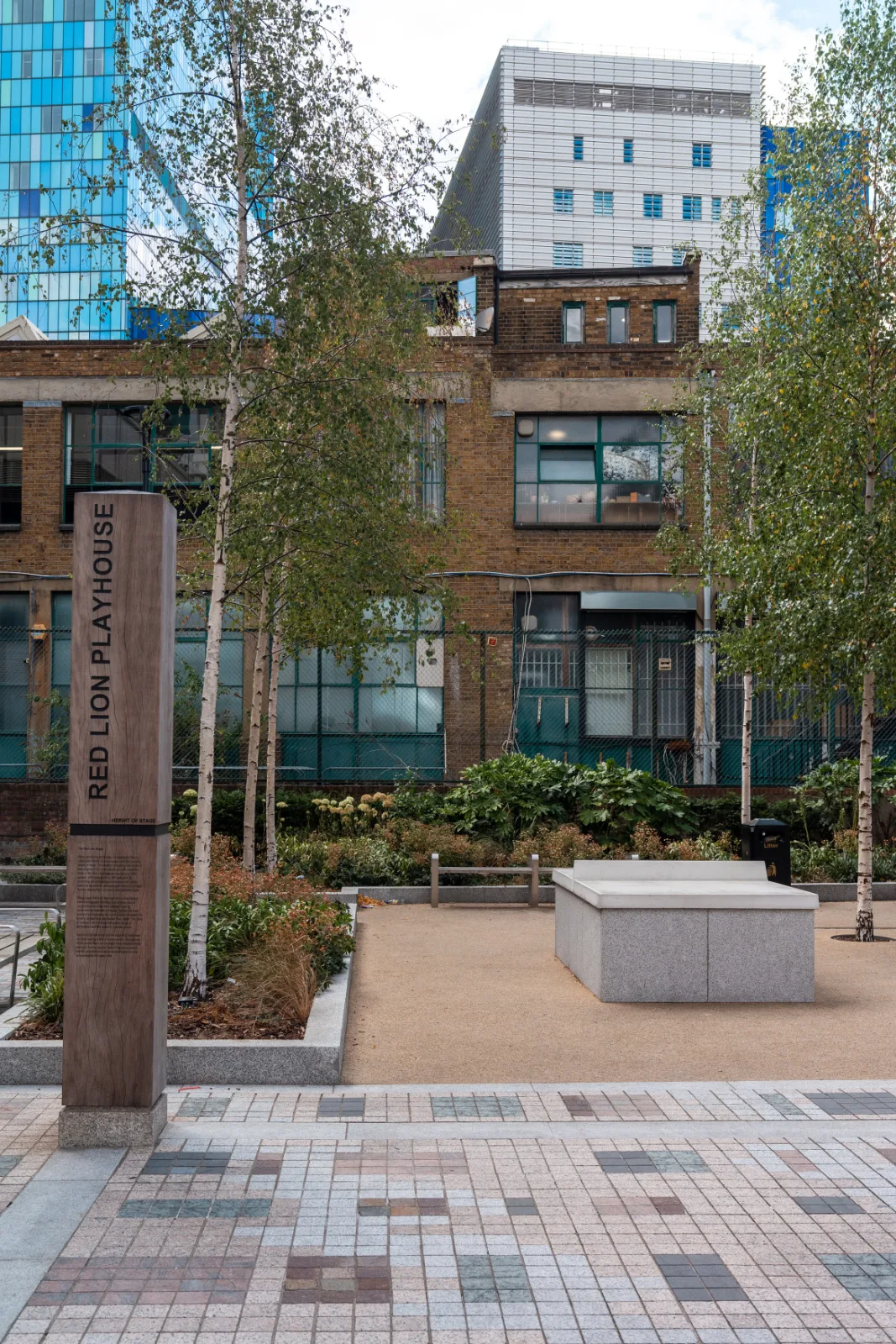A marker post reading Red Lion Playhouse, Height of Stage, standing in a new public square with birch trees and glass towers behind