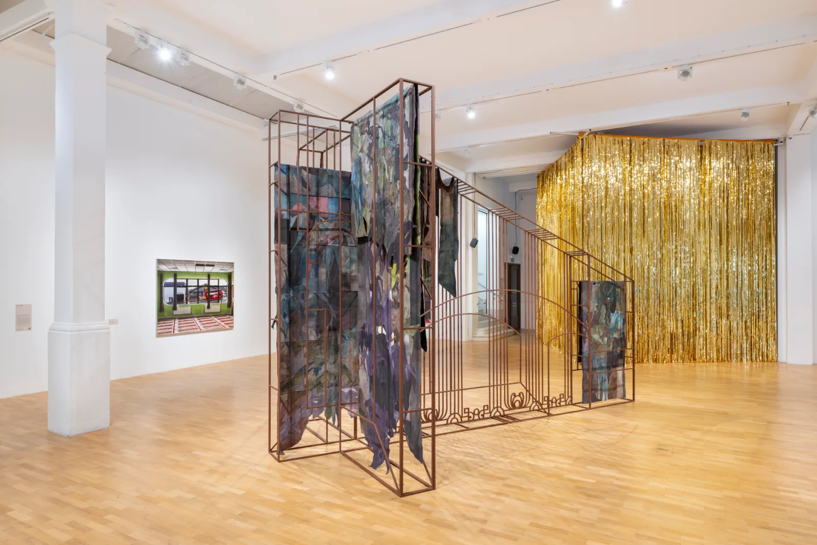 Installation view of East of the Aldgate Pump at the Whitechapel Gallery, with hung textiles on a rusted metal frame and a gold curtain beyond