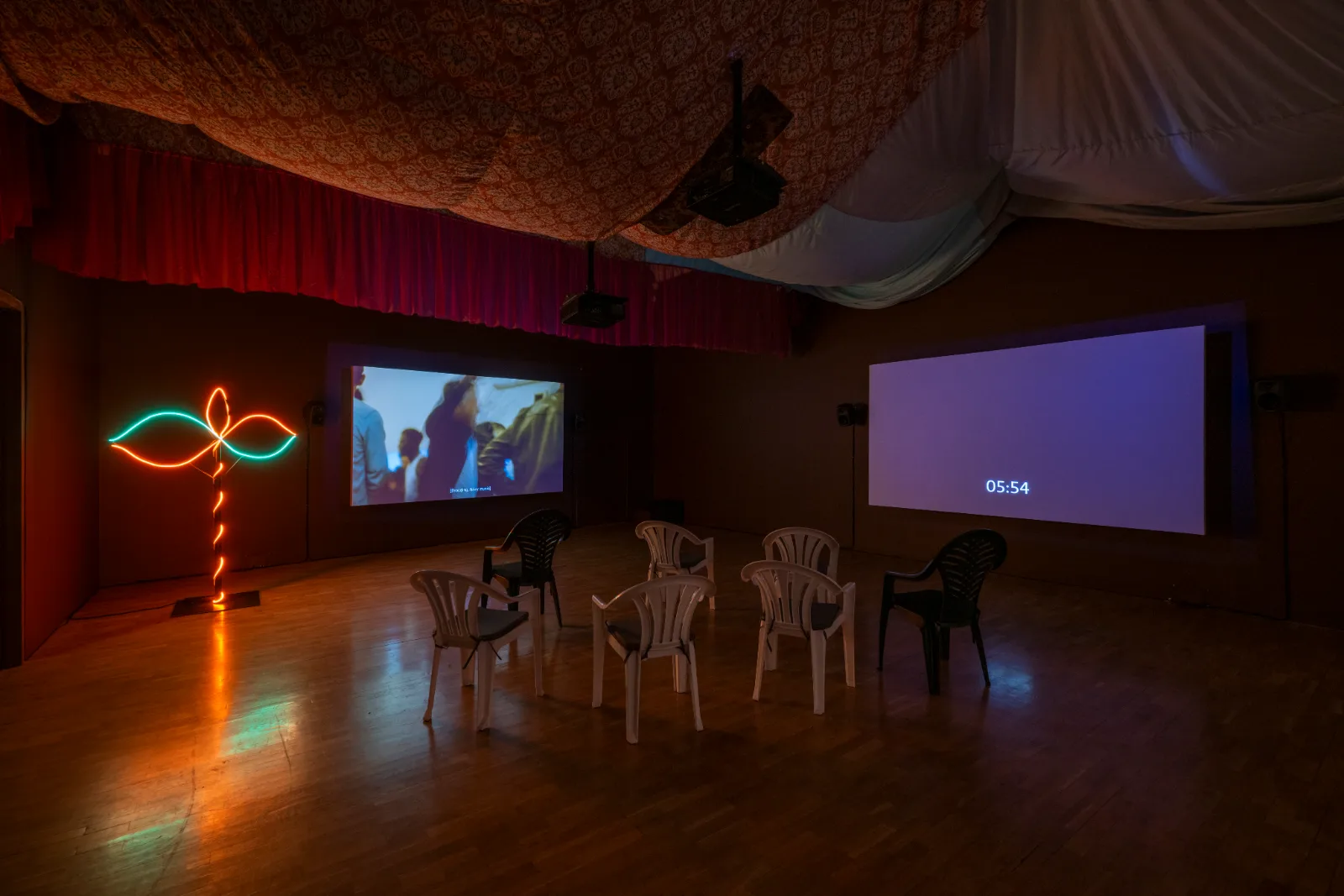 Installation view of East of the Aldgate Pump at the Whitechapel Gallery: a darkened room with two projections, a neon sculpture and plastic chairs beneath a canopy of patterned fabric